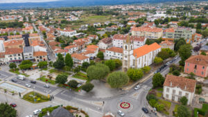 Aerial view of a European town with red-tiled roofs, white church with a tall steeple, and a circular plaza surrounded by streets and greenery.