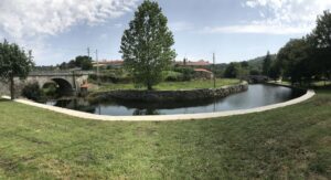 Park scene with a curved waterway, stone arch bridge on the left, and a large tree centered by grassy banks in sunny weather.