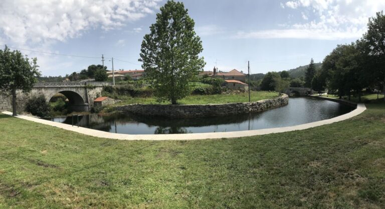 Park scene with a curved waterway, stone arch bridge on the left, and a large tree centered by grassy banks in sunny weather.