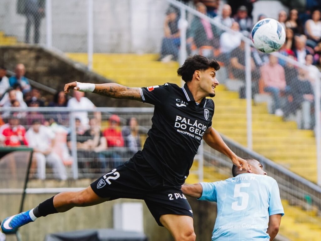 Soccer player in a black kit jumps mid-air to challenge the ball, with an opposing player in light blue behind and a crowded stadium in the background. Académico de Viseu marca passo com nulo frente ao Vizela