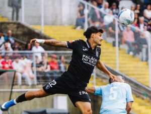Soccer player in a black kit jumps mid-air to challenge the ball, with an opposing player in light blue behind and a crowded stadium in the background.