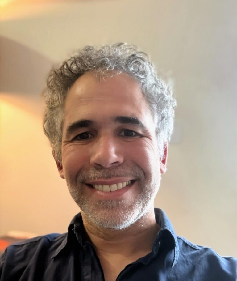 Smiling man with short gray hair and stubble in a dark blue shirt, looking at the camera indoors.