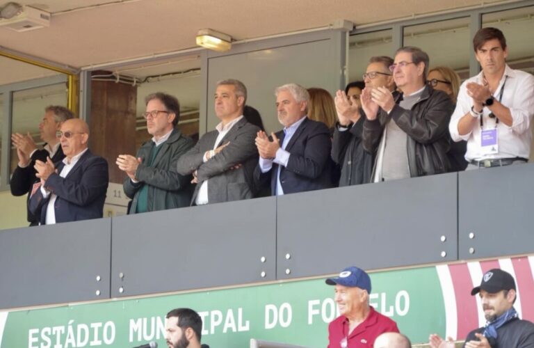 Group of men in suits and jackets standing on a stadium balcony, clapping during an event.