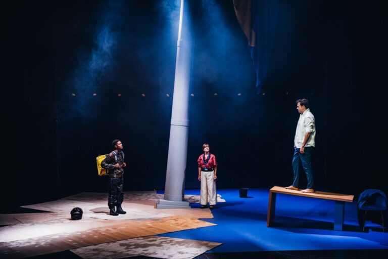 Theater scene with three actors under blue stage lighting: a tall central pillar, left actor with a yellow backpack, center actor, and right actor standing on a bench behind a desk-like surface.