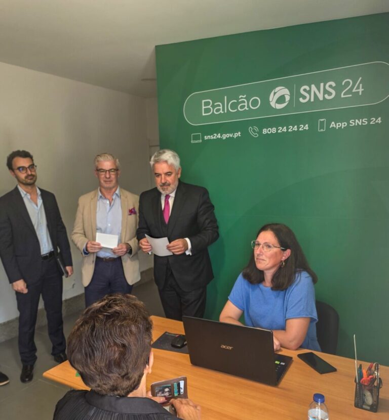 Office scene with a woman at a desk with a laptop, while three men in suits stand with papers in front of a green SNS24 Balcão banner.