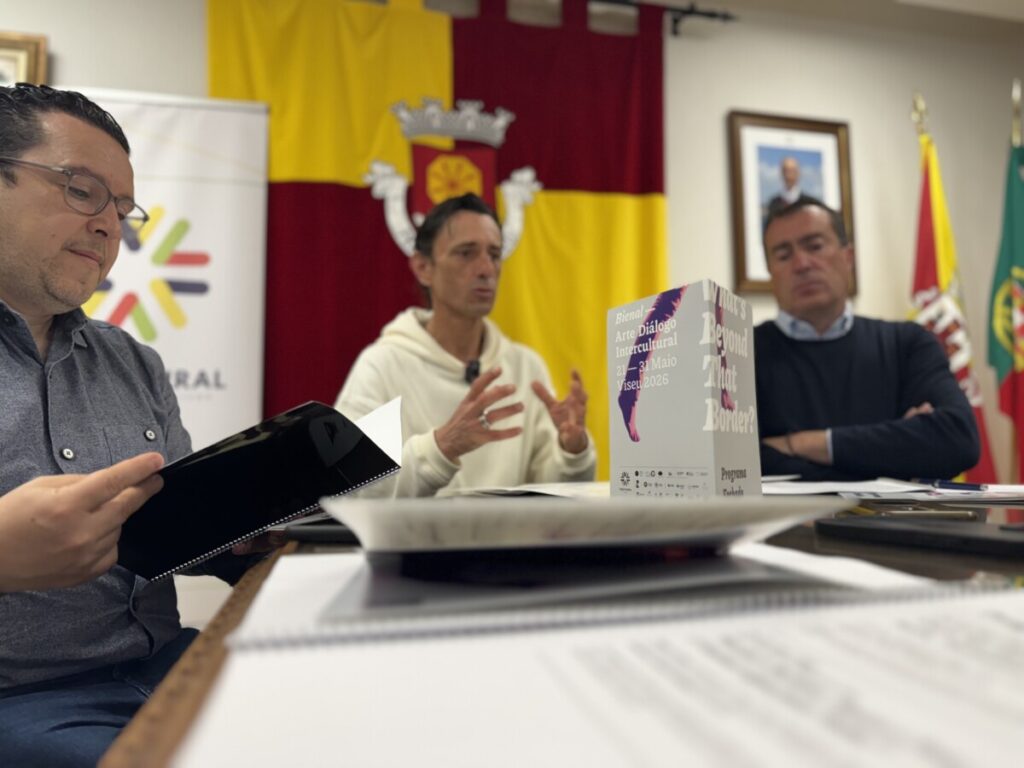 Panel discussion at an intercultural art biennial, with three men seated at a table and flags in the background.