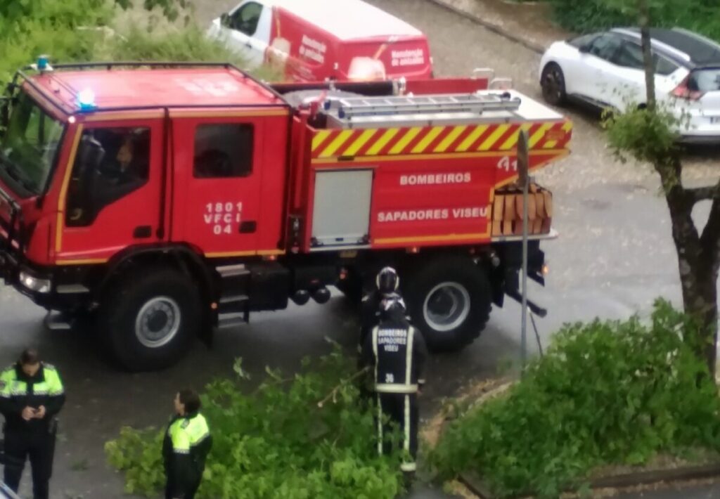 Red fire truck with yellow chevrons parked on a street; firefighters in high-visibility jackets nearby. Mau tempo provoca estragos em Viseu com árvores caídas e estruturas danificadas