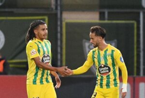 Two soccer players in yellow-and-green striped jerseys shake hands on a football pitch after a play or at a break.