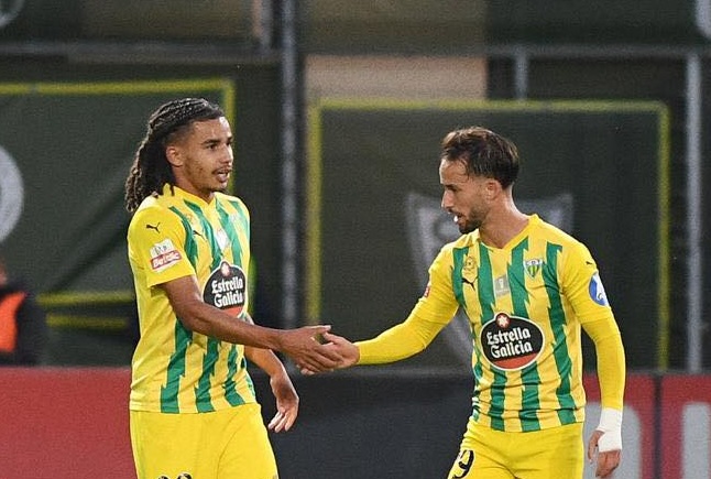 Two soccer players in yellow-and-green striped jerseys shake hands on a football pitch after a play or at a break.