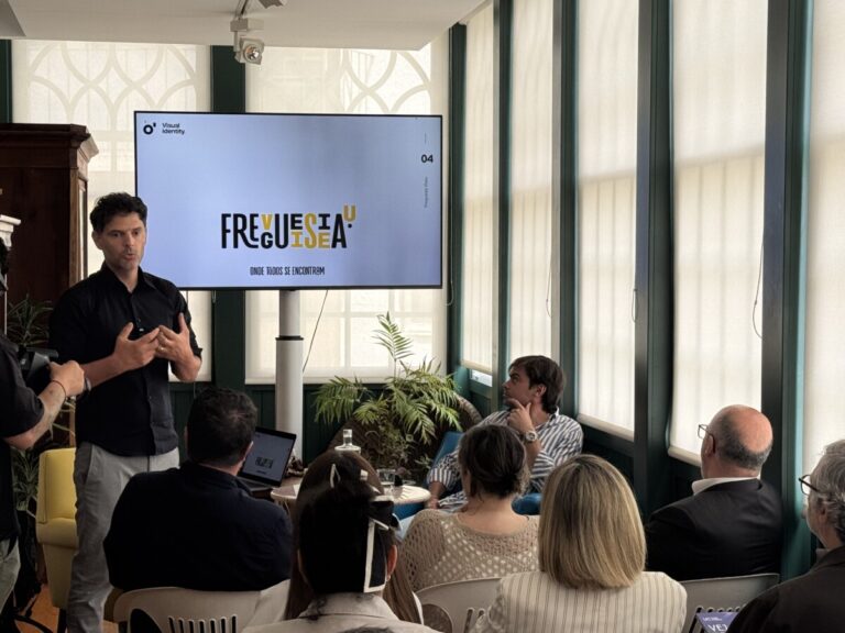 Presenter speaking to an audience in a room with a large screen displaying a logo and text, during a talk or workshop.