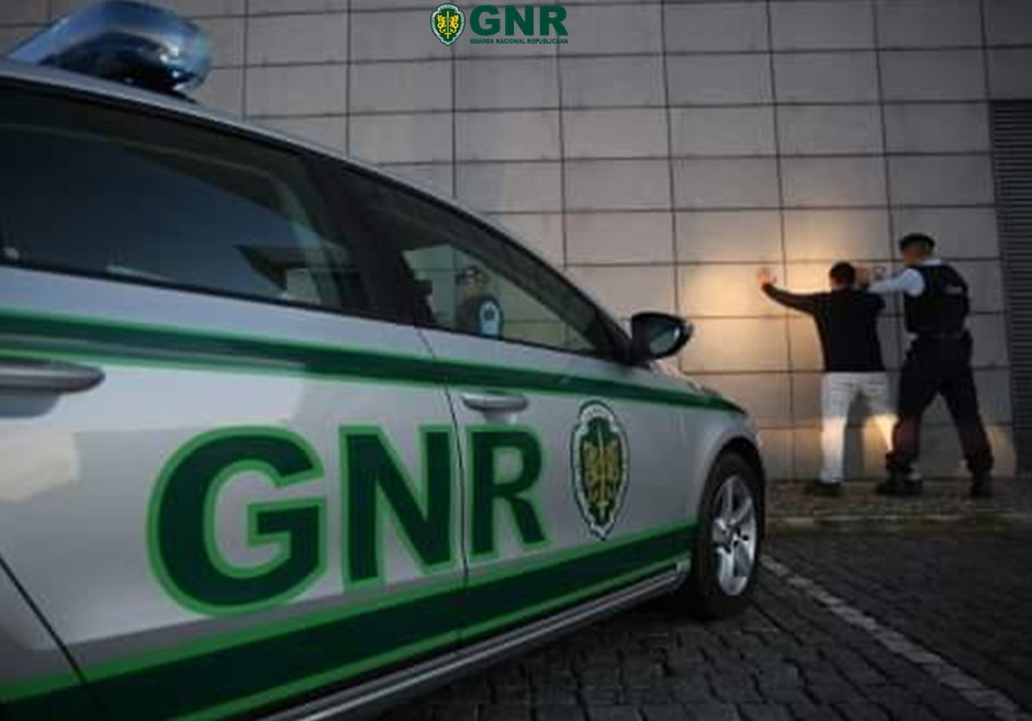 GNR police car in the foreground with two officers restraining a man against a wall. Homem de 35 anos detido pela GNR por furto em residência