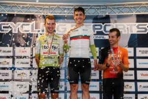 Three male cyclists stand on a podium with medals and confetti, posing for a photo against a cycling event backdrop.