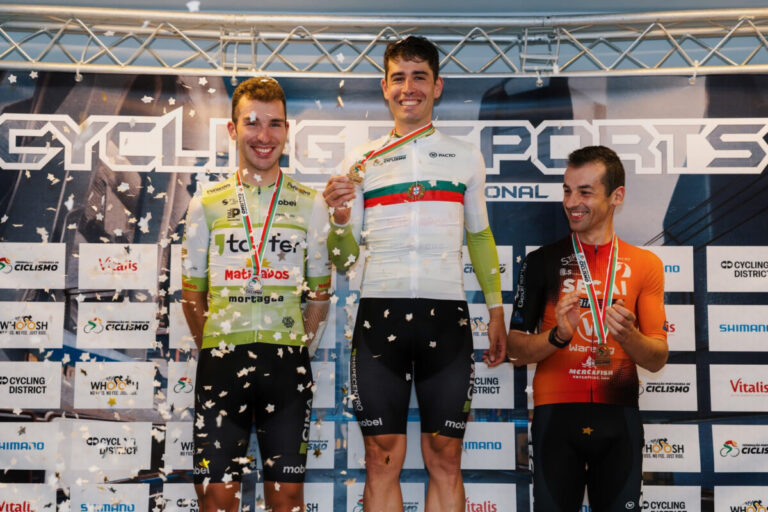 Three male cyclists stand on a podium with medals and confetti, posing for a photo against a cycling event backdrop.