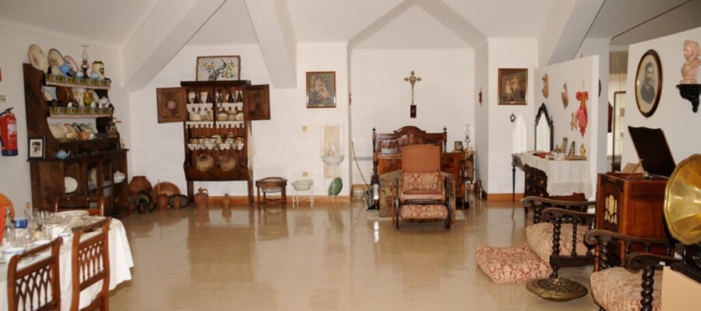 Antique-filled parlor with a central carved bed/chair, upholstered seating, a china cabinet, pottery on shelves, and a brass gramophone on the right. Museu de Silgueiros reabre ao público e celebra património etnográfico de referência nacional
