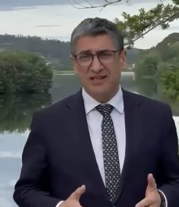 Man in a dark suit, white shirt, and patterned tie speaks to camera outdoors, gesturing with hands, with trees and water in the background.