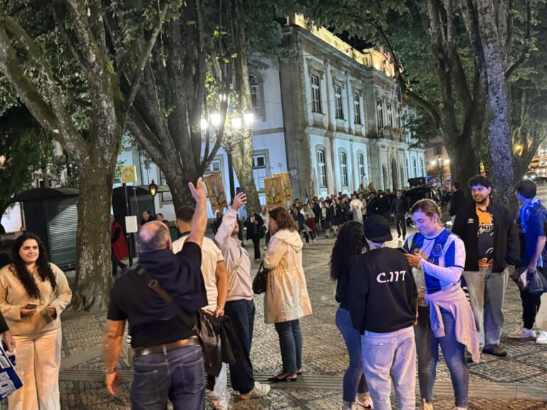 Nighttime street scene of a crowd walking in a procession along a cobblestone street, flanked by large trees and a blue historic building with lit windows and banners.