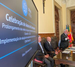 Man in a suit speaks at a formal meeting; three panelists sit beside him in an ornate room with flags and a large screen displaying the event text on the left.