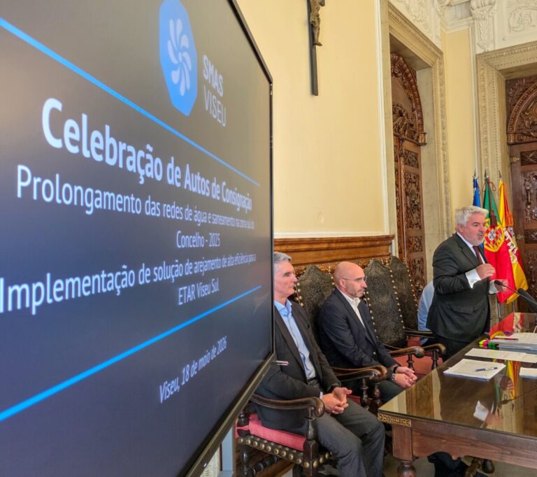Man in a suit speaks at a formal meeting; three panelists sit beside him in an ornate room with flags and a large screen displaying the event text on the left.