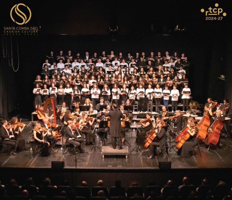 Full orchestra and large choir performing on a dimly lit stage, conductor center with musicians and audience in foreground; logos in top corners.