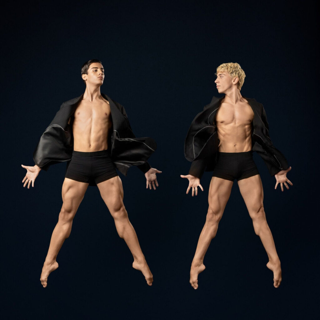 Two male dancers leap mid-air in black jackets and shorts against a dark backdrop, arms extended wide.