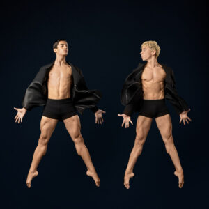 Two male dancers leap mid-air in black jackets and shorts against a dark backdrop, arms extended wide.