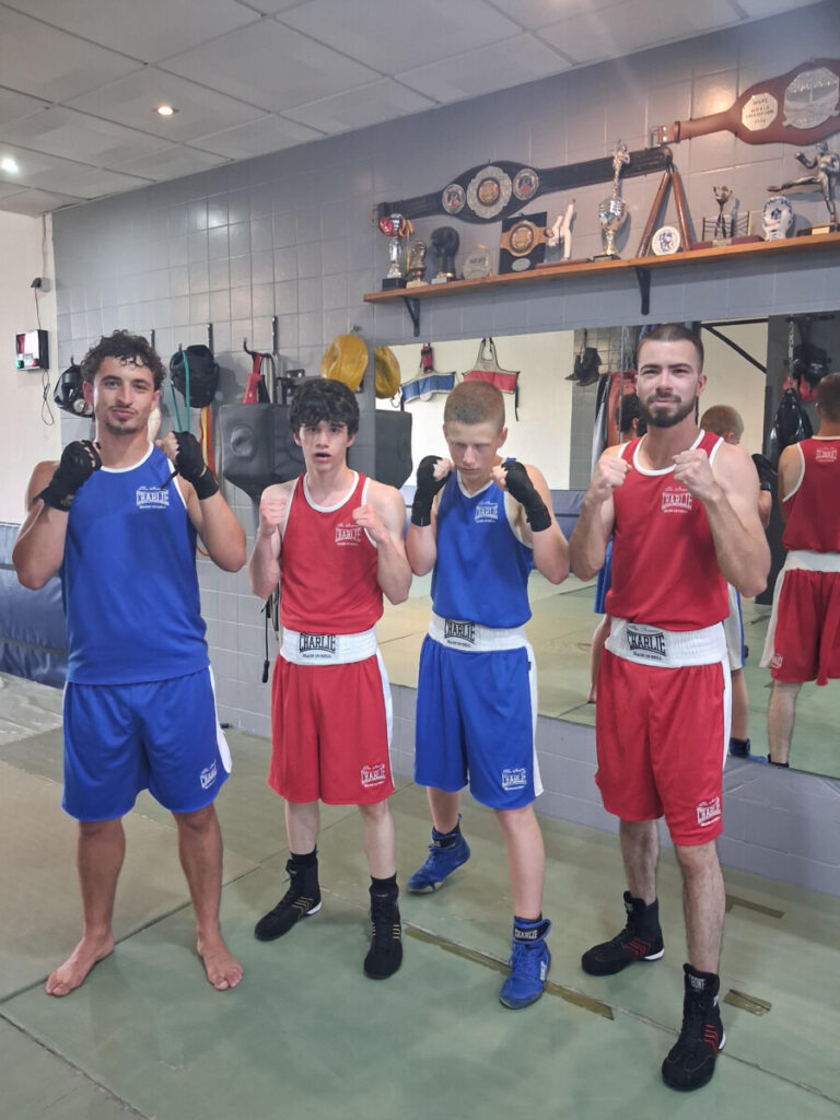 Four young boxers in a gym pose with fists up, wearing red and blue uniforms, trophies on a shelf in the background.