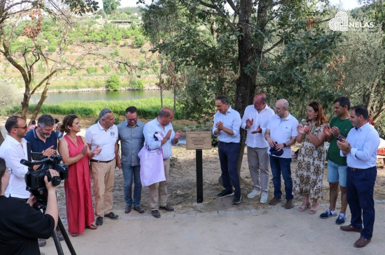 Group of adults in casual and formal clothing standing in a circle outdoors, unveiling a plaque under a tree beside a river.