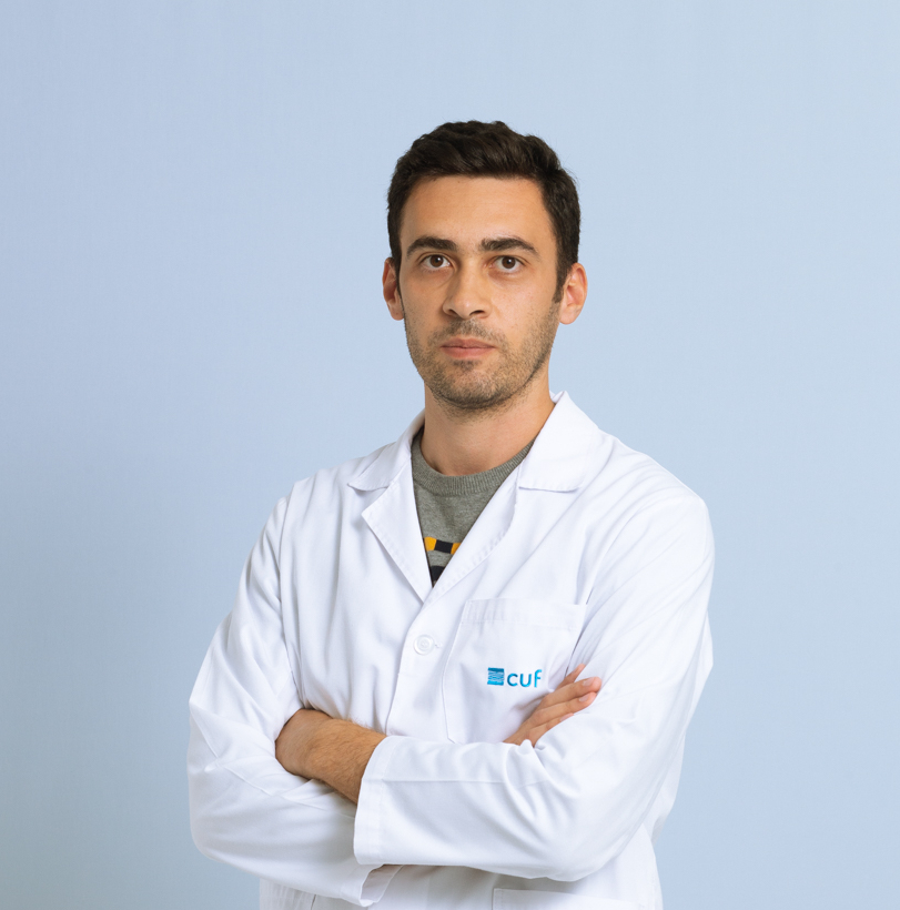 Medical professional in a white lab coat with arms crossed, standing against a light blue background.