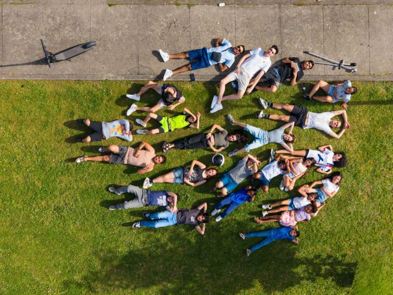 Aerial view of a diverse group of people lying on green grass, arranged to form a heart shape from above, with a paved path at the top.