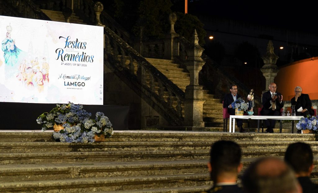 Nighttime outdoor stage with three men seated at a table, microphones, and blue floral arrangements; a large banner reading 'Festas Remédios' on the left. Xutos & Pontapés e Plutónio na “romaria” de Lamego