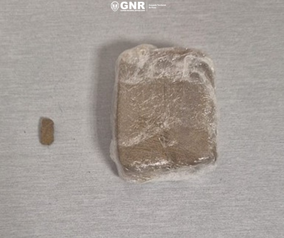 Brown rectangular block wrapped in plastic on a light surface, with a small piece to its left. GNR detém dois homens por tráfico de droga em ação de fiscalização rodoviária em Viseu