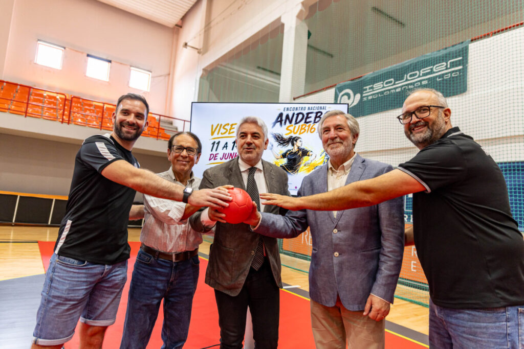Five men in casual and business attire stand on a gym floor, holding a red ball together and smiling nearby bleachers. Arranca esta quinta-feira em Viseu a fase final do Campeonato Nacional de Andebol Feminino Sub-16