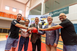 Five men in casual and business attire stand on a gym floor, holding a red ball together and smiling nearby bleachers.