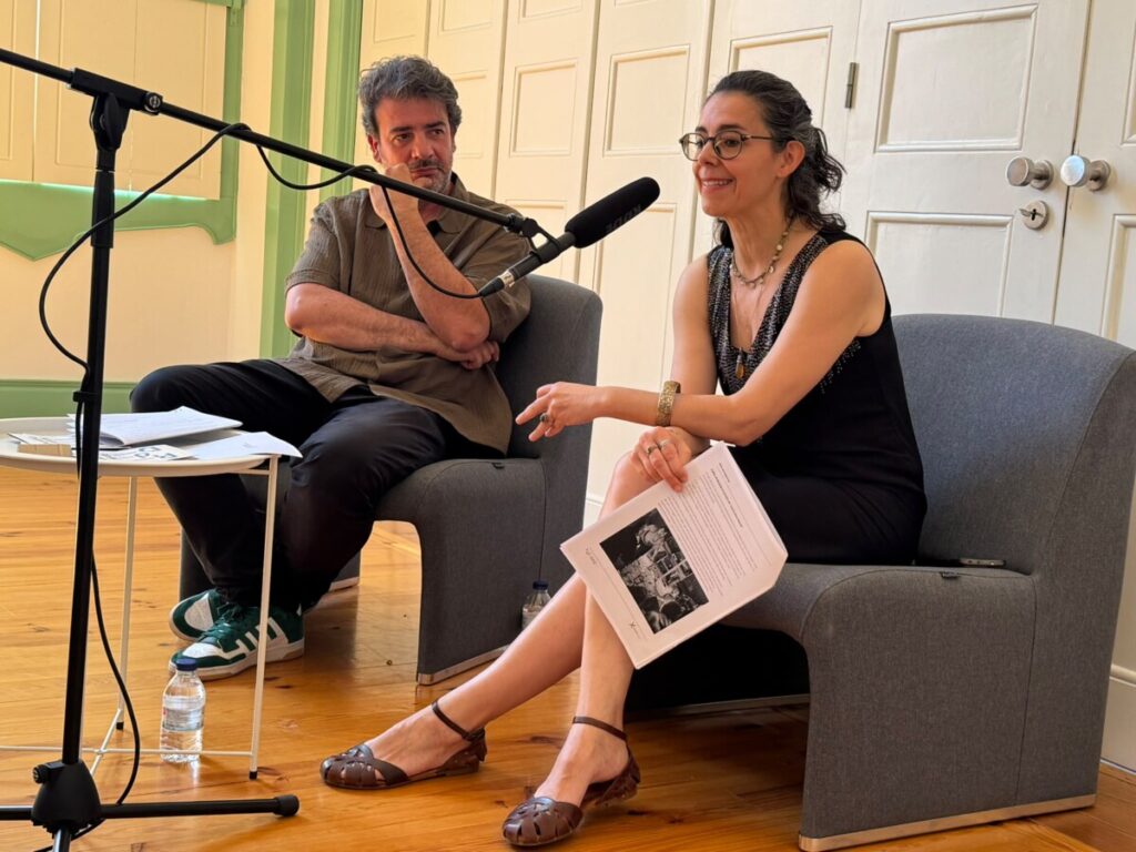 Two people in a studio speak into a/ their microphone setup during a talk or podcast recording, seated on gray chairs with papers in hand.