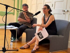 Two people in a studio speak into a/ their microphone setup during a talk or podcast recording, seated on gray chairs with papers in hand.