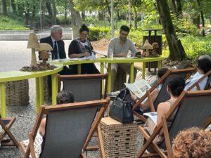 Outdoor panel discussion in a park with three speakers at a lime-green table and an audience seated in wooden lounge chairs.