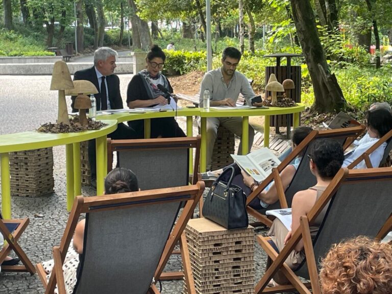 Outdoor panel discussion in a park with three speakers at a lime-green table and an audience seated in wooden lounge chairs.