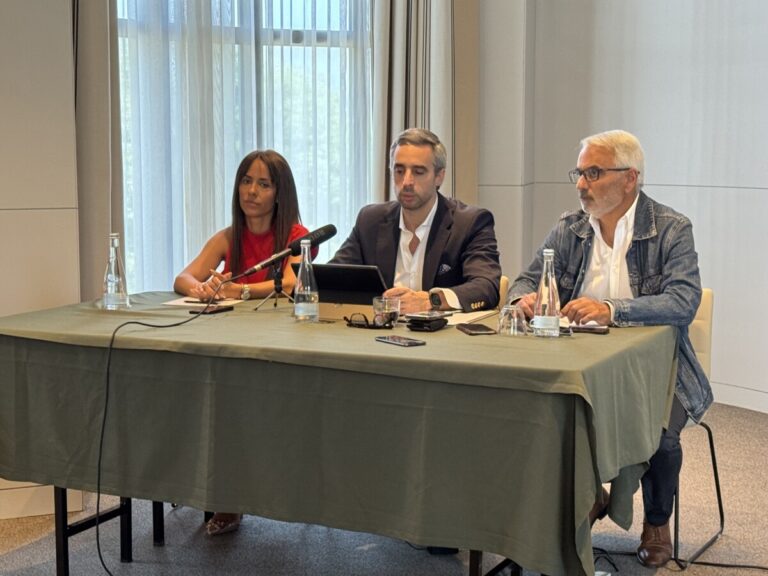 Three adults seated at a conference table with microphones, speaking at a panel briefing.