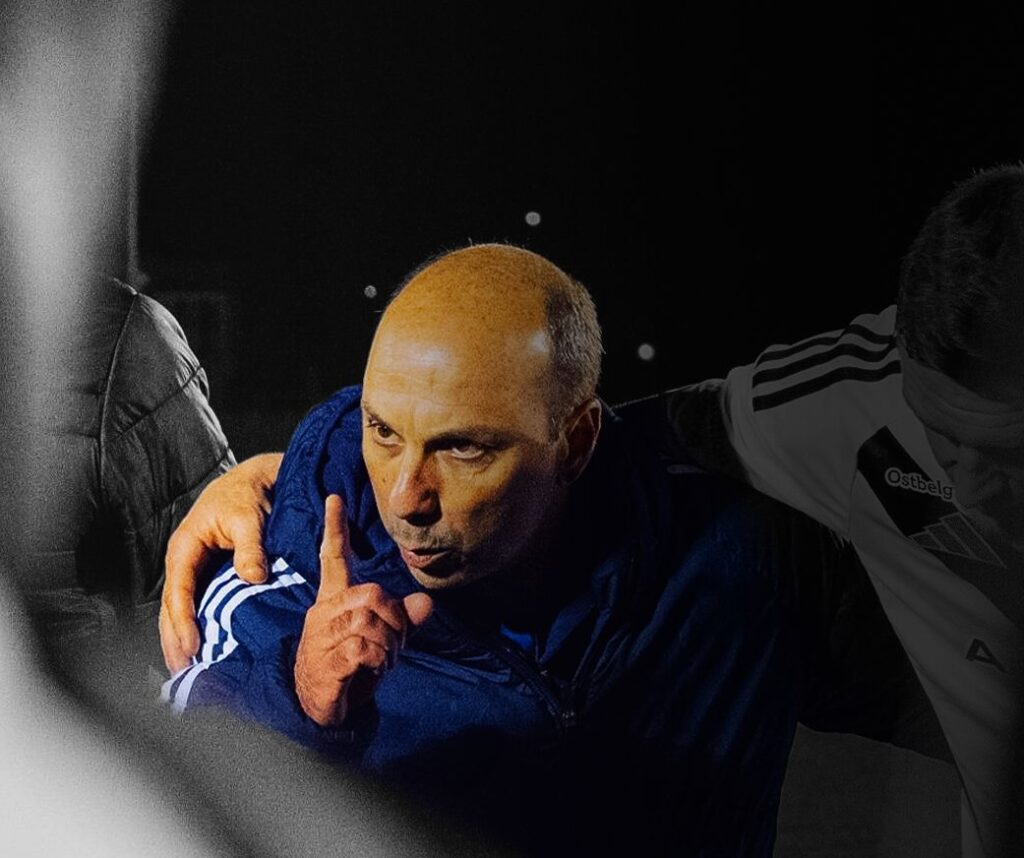 Coach in a blue jacket holds a teammate, raising a finger to his lips as a signal to be quiet during a night practice or game show of focus. Bruno Pinheiro é o treinador do Académico de Viseu