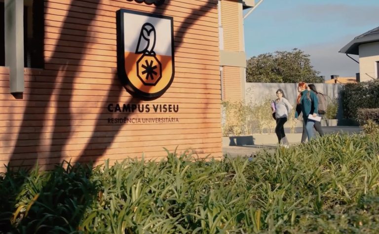 Campus Viseu university residence sign on a brick wall with a shield logo, greenery in front, and students walking by.