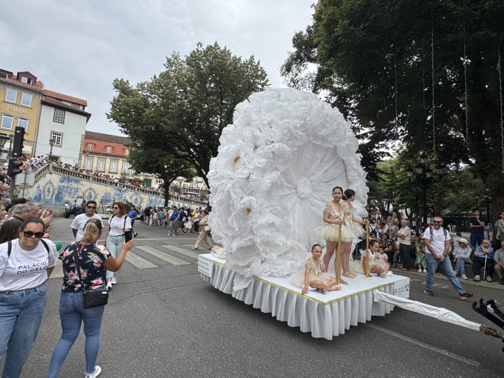 Giant white flower float with four young dancers in cream tutus standing and kneeling on a parade platform, crowds watching along a city street.
