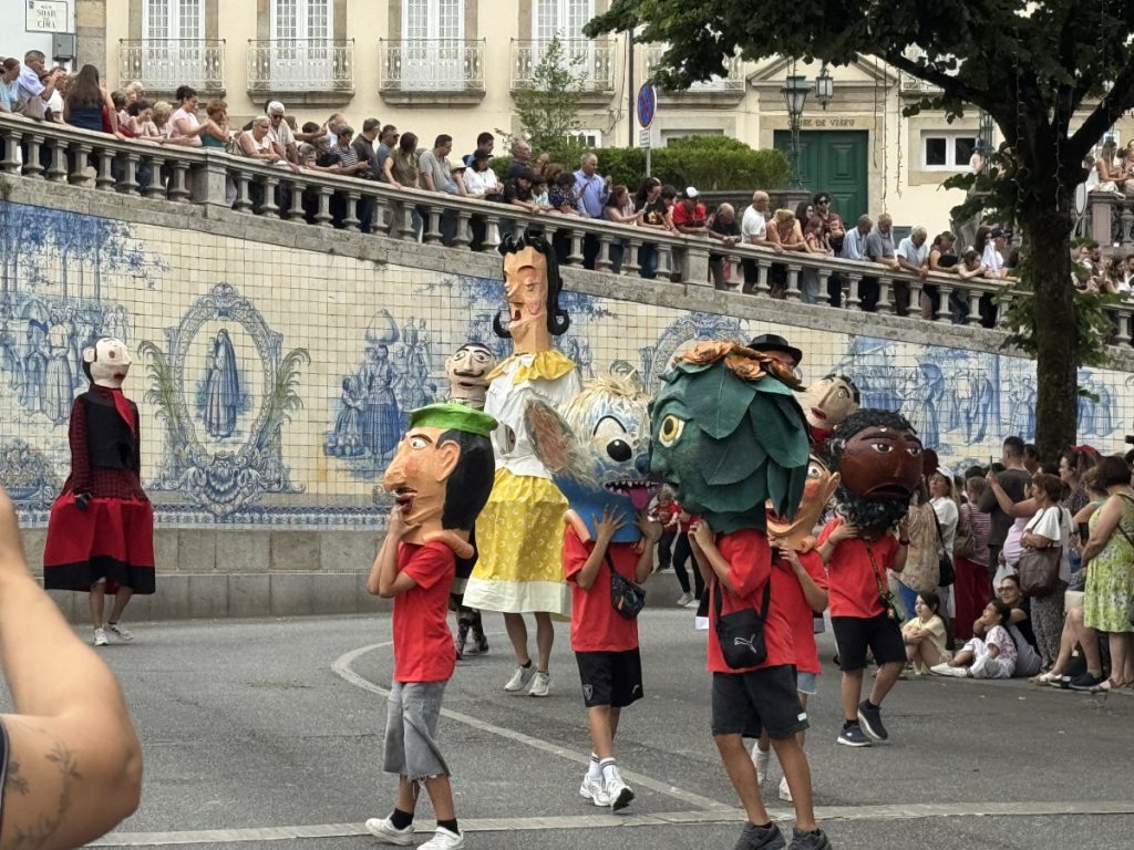 Children in red shirts carry large colorful puppet heads in a city street parade as spectators line a tiled mural wall behind them.