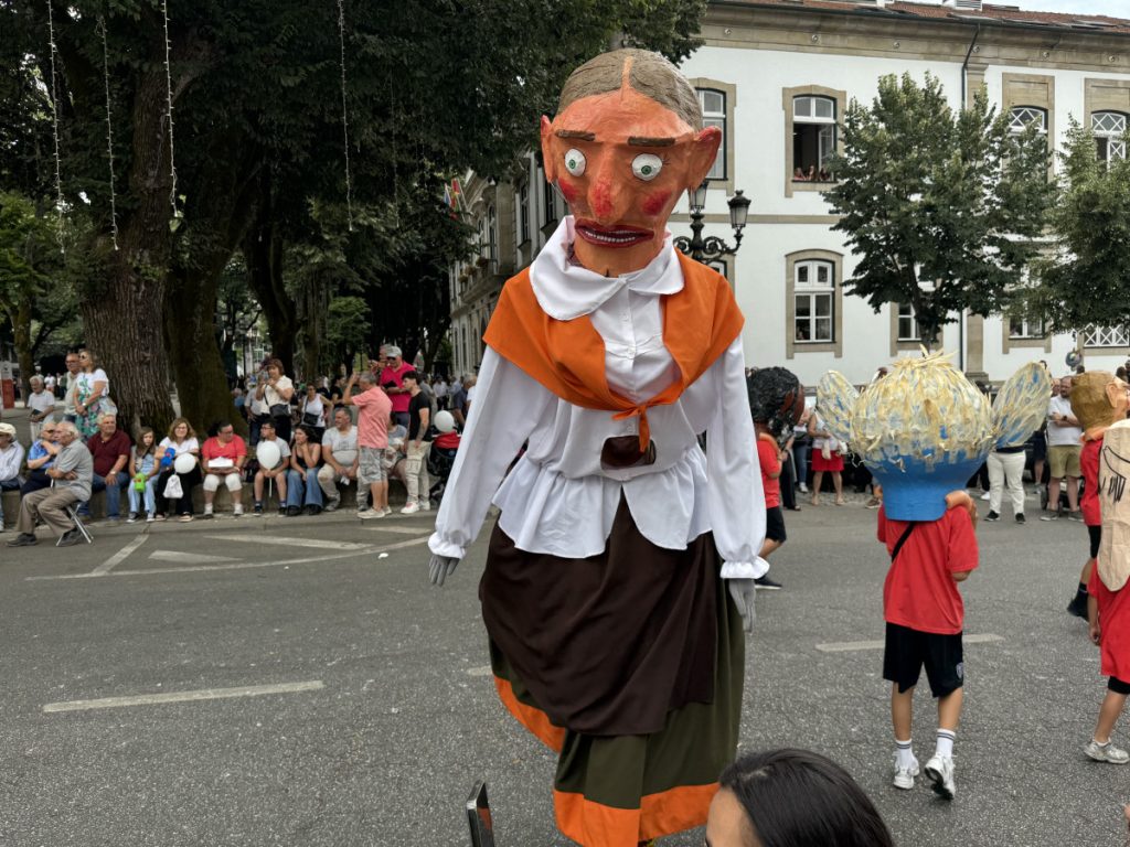 Giant puppeted figure in an orange shawl and white blouse parades down a city street as spectators watch from the curb.