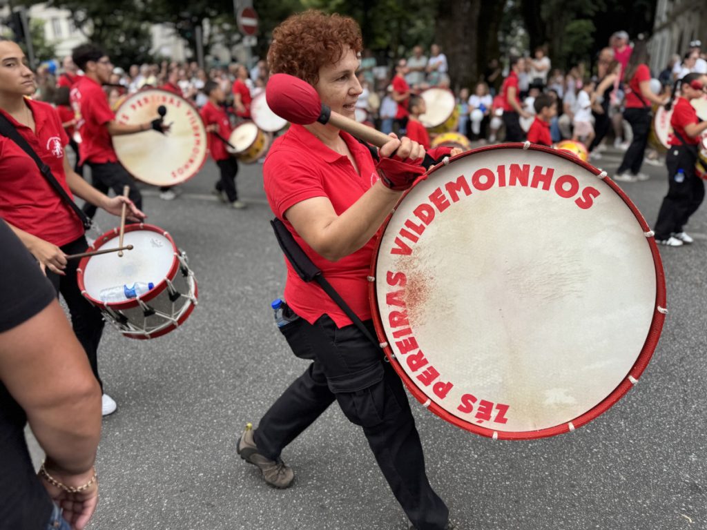 Drummer in a red shirt leads a street parade, playing a large bass drum close to the camera.