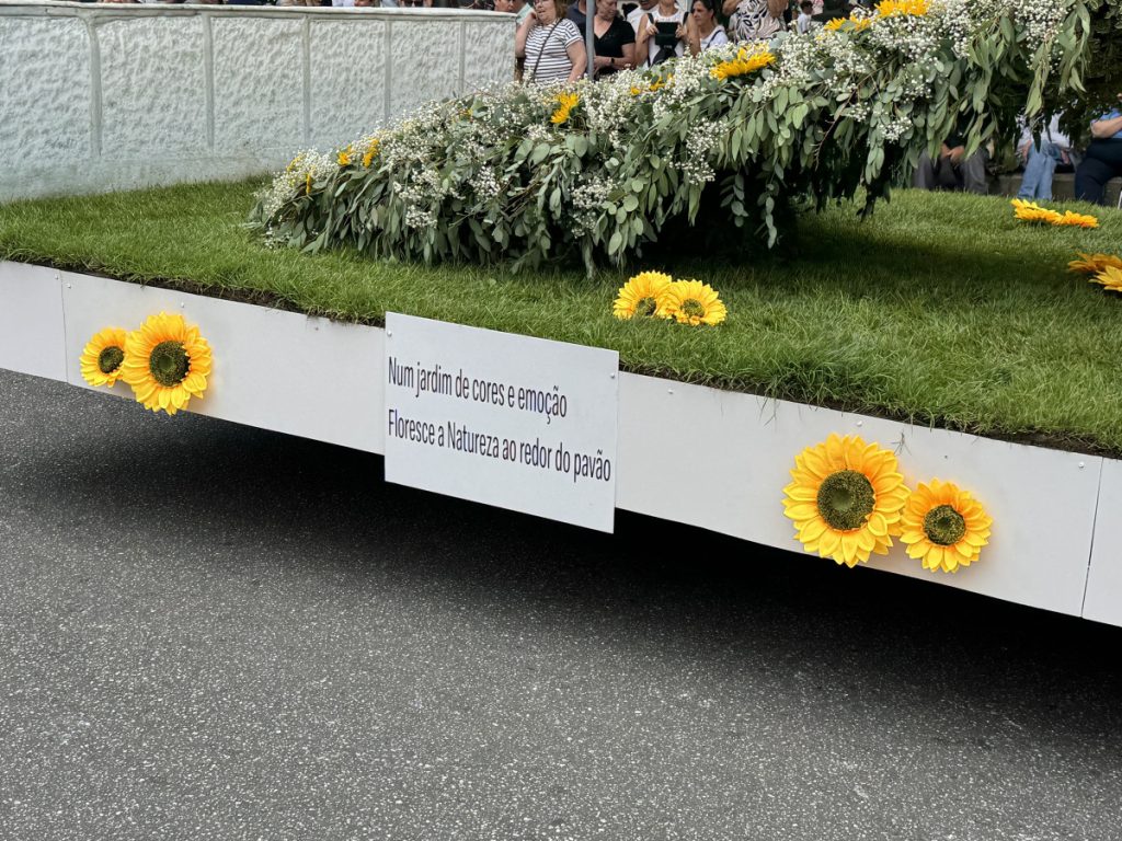 Floral parade float with a grassy garden bed, bright sunflowers, and a white sign bearing Portuguese text, spectators visible in the background.