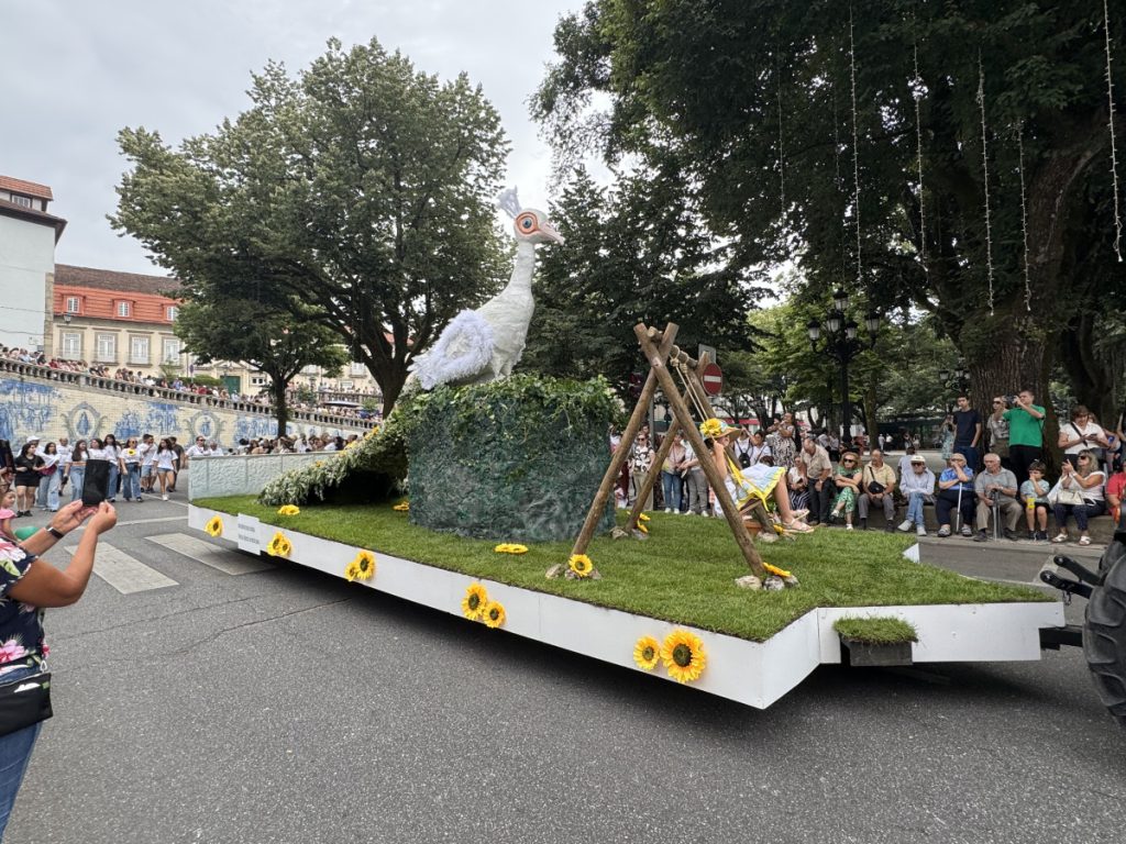 Parade float featuring a large white bird statue on a grassy mound covered in ivy, surrounded by sunflowers, as spectators line the street.