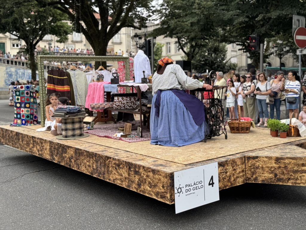 Parade float with a woman in period clothing sewing at a vintage sewing machine while spectators watch behind.