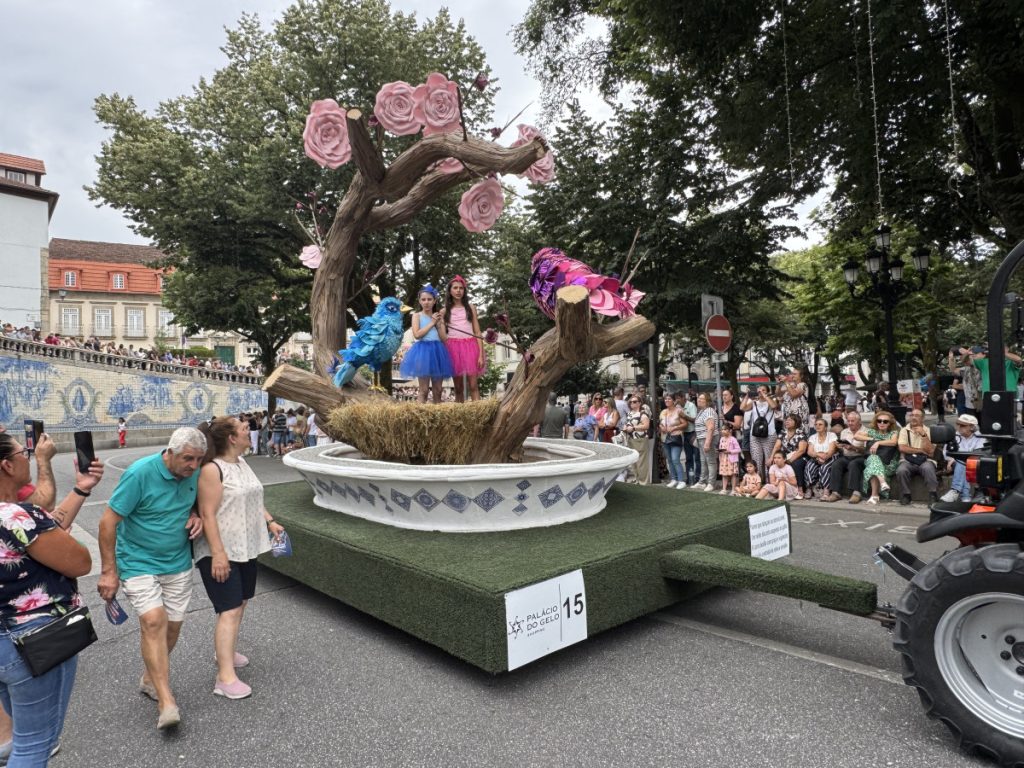 Parade float featuring a twisted tree with pink rose sculptures, two girls in blue and pink tutus perched on the branches inside a large decorative planter; spectators line the street.