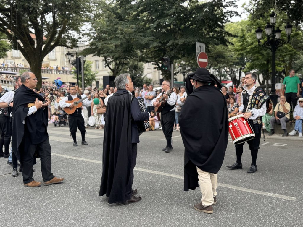 Street performers in dark capes playing guitars, an accordion, and drums during an outdoor festival in front of a cheering crowd beneath trees and historic buildings.