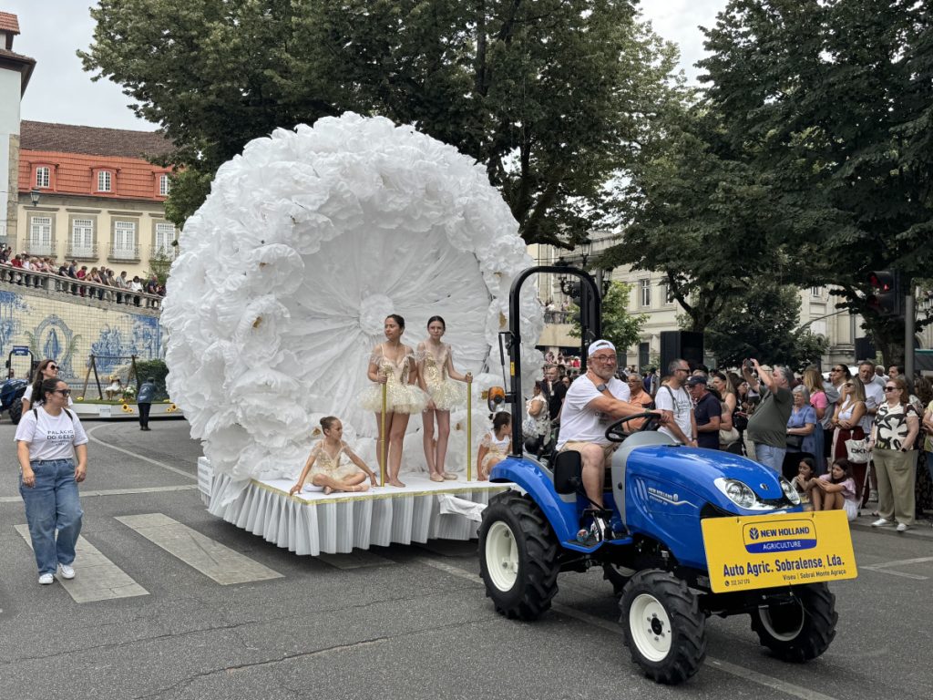 Blue tractor pulls a parade float shaped like a giant white flower, with four young ballet dancers in cream costumes on a pedestal.